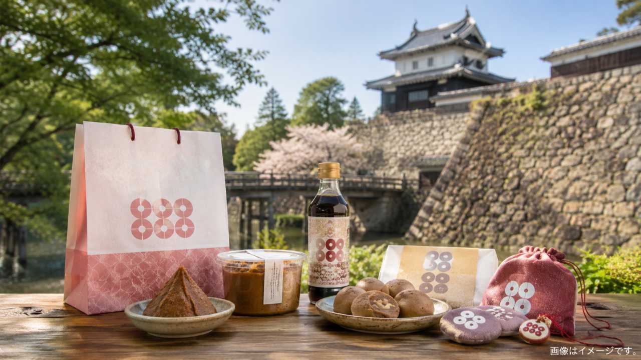 上田城跡公園の櫓と石垣を背景に、和紙風の土産袋、くるみ菓子、味噌、美味だれ風の瓶、六文銭モチーフの小物